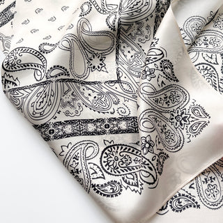 Light beige silk scarf with intricate black paisley pattern, featuring teardrop designs and floral details with a subtle sheen characteristic of premium silk
