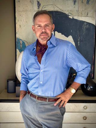 Man wearing purple paisley silk neckerchief with blue striped shirt and grey pants, styled in front of a painting for a classic, refined look.