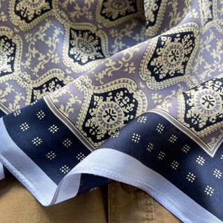 Luxurious silk scarf with ornate geometric floral pattern in navy blue and lilac with cream accents, featuring white border and shiny fabric texture