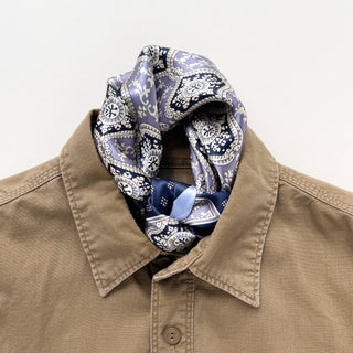 Silk bandana in navy and lavender paisley pattern, elegantly knotted at the neck of a khaki button-up shirt, showcasing versatile styling.