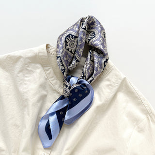 Navy and light blue silk scarf with geometric floral pattern elegantly tied around collar of cream shirt, displaying luxurious sheen and intricate design