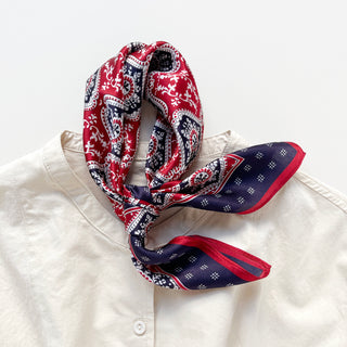 Red and navy silk bandana with geometric floral pattern, elegantly tied around white shirt collar, showcasing intricate paisley design.