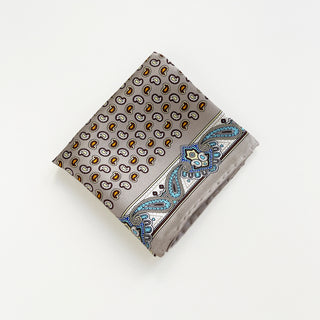 Folded taupe silk scarf with small paisley print pattern in gold and white, featuring decorative blue paisley border with hand-rolled edges