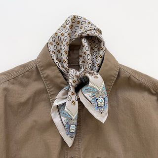 Taupe silk scarf with paisley pattern in blue, gold and brown tones, styled as a neckerchief around the collar of a beige linen shirt