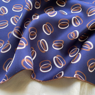 Indigo purple silk scarf with copper and white orbital pattern, showing luxurious twill texture and hand-rolled edge detail on cream surface
