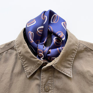 Indigo purple silk scarf with copper and white orbit pattern styled as a neckerchief with a beige button-up shirt, showing luxurious texture and hand-rolled edges.