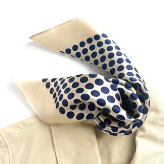 Square silk scarf with navy blue polka dots on light beige background, elegantly draped to showcase its sheen and pattern