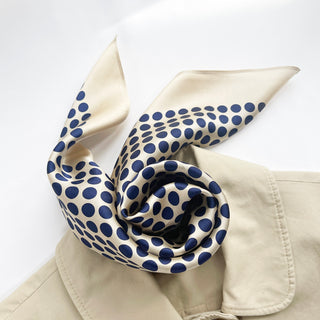 Elegant silk bandana scarf with navy blue polka dots on cream background, displayed with gentle folds showing its lustrous texture