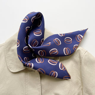 Indigo purple silk scarf with circular orbit pattern in copper and white, elegantly tied on beige jacket collar, showcasing hand-rolled edges and luxurious texture