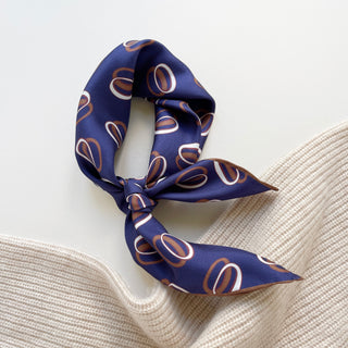 Indigo purple silk bandana with modern orbit pattern in copper and white, tied elegantly on cream knit fabric, showing luxurious shine and hand-rolled edges