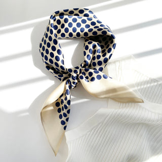 Silky square bandana scarf with navy blue polka dots on cream background, elegantly tied in classic neckerchief style, displayed over white fabric.