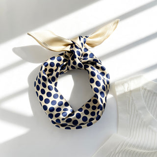 Cream silk bandana scarf with navy blue polka dots, tied in a stylish knot on white surface with soft shadows