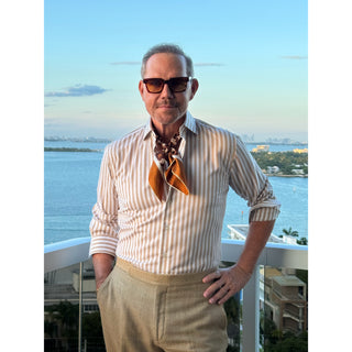 Man wearing a brown tones silk square neckerchief featuring retro geometric pattern in a stylish outfit standing on a balcony with a scenic view of water and buildings