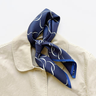 Navy blue silk scarf with white chain-link pattern and dotted design, featuring royal blue edges, displayed elegantly on beige jacket collar