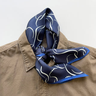 Navy blue silk neckerchief with white geometric pattern and blue trim, elegantly knotted at the collar of a beige shirt