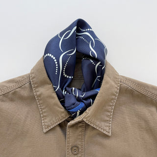 Navy blue silk neckerchief with white geometric pattern and dotted lines, elegantly tied at the neck of a beige linen shirt