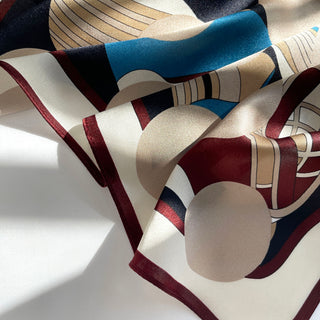 Silk Bauhaus-inspired scarf with geometric patterns in burgundy, blue, beige, and navy, featuring a lustrous sheen and bordered edge