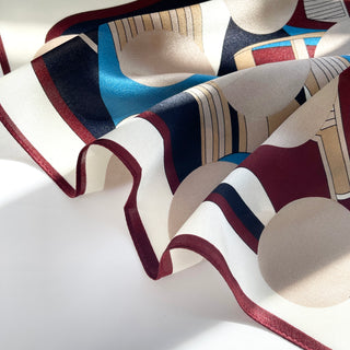 Silk scarf with Bauhaus-inspired geometric pattern featuring burgundy, blue, beige, and black shapes on white background with burgundy edge trim