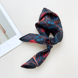 Luxury silk bandana scarf with navy blue background featuring vibrant red and blue floral patterns, shown folded with hand-rolled edges displayed on white surface.