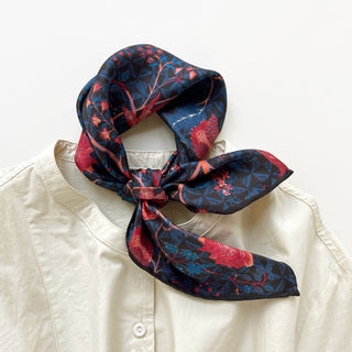 Midnight blue silk bandana scarf with vibrant red floral design, displayed tied around collar of cream shirt, showing hand-rolled edges and luxurious twill texture.