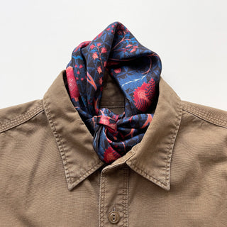 Navy silk bandana scarf with vibrant red floral pattern elegantly tied at the neck of a tan button-up shirt, showcasing luxurious hand-rolled edges