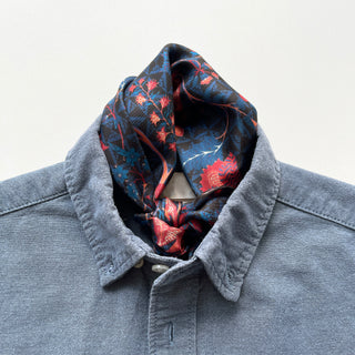 Navy silk scarf with red floral pattern styled as neckerchief with chambray button-up shirt, showing luxurious hand-rolled edges and twill texture.
