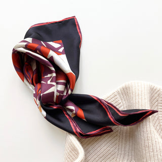 Luxury silk bandana with burgundy, purple, and red berry blossom pattern, featuring hand-rolled edges and navy border, draped elegantly on cream knit fabric
