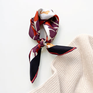 Silk bandana tied in knot displaying burgundy, red, gold and white floral pattern with hand-rolled edges beside cream knit fabric