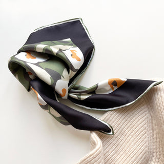 Luxury silk scarf with abstract floral pattern in deep purple, green, white, and orange accents, featuring hand-rolled edges on white background