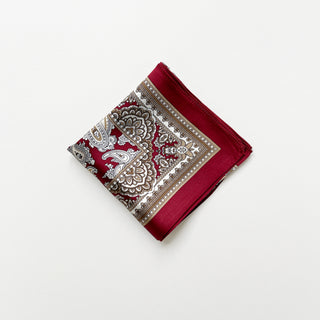 Folded dark red silk neckerchief featuring intricate beige paisley patterns and geometric border details on a white background