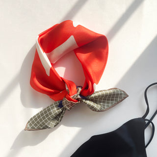 Vibrant orangey-red silk scarf with white accents tied in a knot, featuring green checkered pattern on the ends, displayed on white surface in natural light
