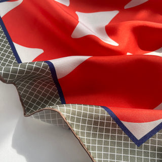 Close-up of vibrant orangey-red silk scarf with white abstract shapes and green checkered pattern edged with navy blue trim, showing luxurious silk texture