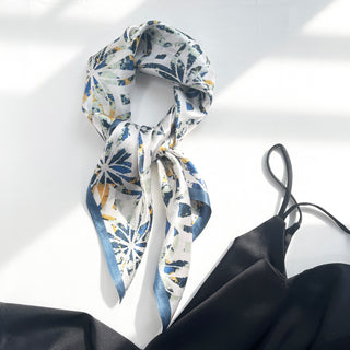 Dusty blue silk bandana scarf with artistic floral pattern in blue and yellow tones, styled in a knot with blue border trim against white background