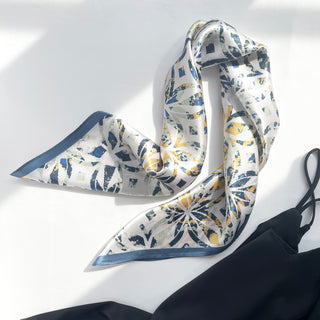 Square silk bandana scarf with abstract dusty blue and yellow floral pattern on white background, featuring blue border trim, displayed elegantly folded