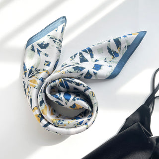 Silk bandana scarf with dusty blue border and abstract floral pattern in blue and yellow, elegantly draped on white surface showing lustrous texture