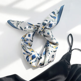 Silk bandana scarf with dusty blue and yellow abstract floral pattern, tied in a stylish knot against white background, displaying silky texture