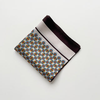 Folded silk bandana scarf with colorful geometric pattern featuring blue, red, and yellow motifs, bordered with burgundy and white stripes