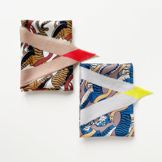 Two folded Tiger Reverie scarves - warm earth tones with red accent on left, cool blue and grey with yellow accent on right, both featuring tiger pattern.