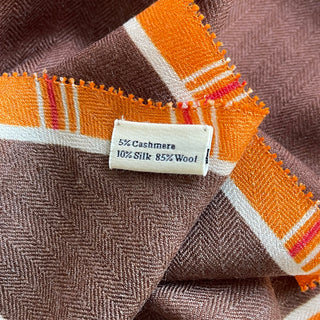 Close-up of brown and orange plaid cashmere-silk scarf showing herringbone texture, orange border with accent stripes, and fabric content label.
