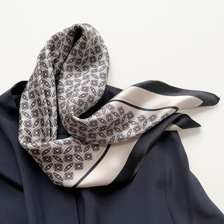 Luxurious silk bandana scarf with black and cream geometric pattern, featuring elegant leaf motifs and bordered design, draped on dark fabric.