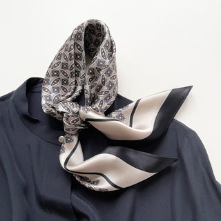 Elegant silk scarf with geometric pattern in silver, black and cream tones, styled around a dark blue shirt collar, showcasing sheen and versatile drape