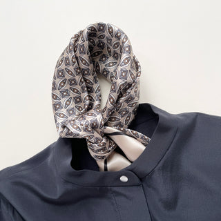 Silk bandana scarf with intricate geometric pattern in black, red and cream tones, elegantly tied around collar of navy blue shirt.