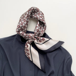 Luxurious silk bandana scarf with red and black geometric heritage pattern tied elegantly around a navy shirt collar, displaying its shiny texture.