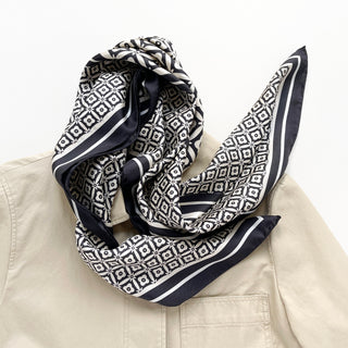 Elegant black and white silk scarf with geometric floral pattern and border, draped artfully on a beige coat, showcasing luxurious texture and hand-rolled edges.