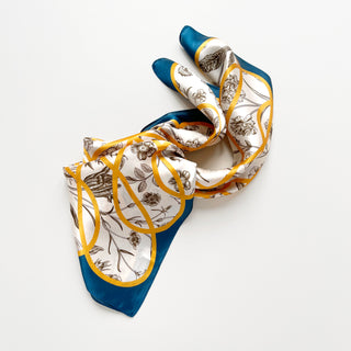 Elegant silk scarf with botanical sketches on ivory background, framed by teal border and gold ribbon details, displayed in gentle folds