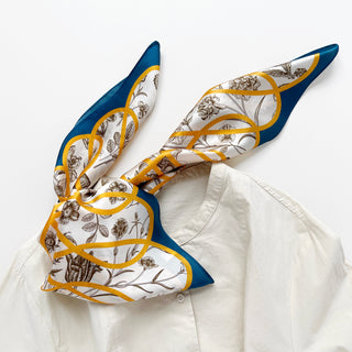 Luxurious silk scarf featuring botanical sketches on ivory background with golden ribbon pattern and teal border, elegantly draped on white fabric