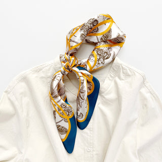 Silk scarf with botanical illustrations and golden ribbon pattern on ivory background with teal border, elegantly tied around shirt collar