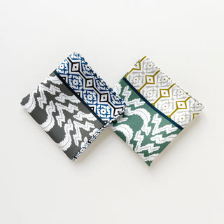 Two folded silk scarves displayed side-by-side - one with gray and blue diamond patterns, and one with teal green and gold geometric designs and hand-rolled edges.
