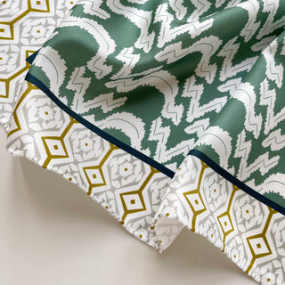 Teal green silk scarf with geometric zigzag pattern and navy blue hand-rolled edge, displayed alongside white and gold diamond pattern design