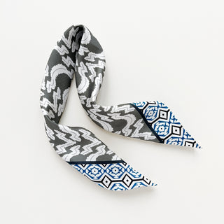 Silk scarf featuring grey and white zigzag pattern with teal blue geometric border design, elegantly twisted to display hand-rolled edges